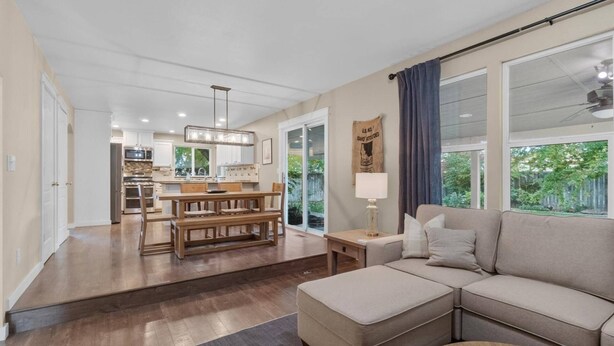 A spacious living room with a large sectional sofa, a wooden dining table with benches, a kitchen in the background, and a view of greenery outside.