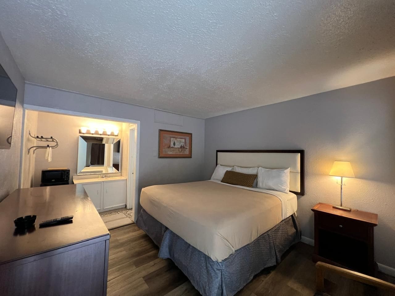 A hotel room with a bed, bedside table, lamp, and a mirror with lights.