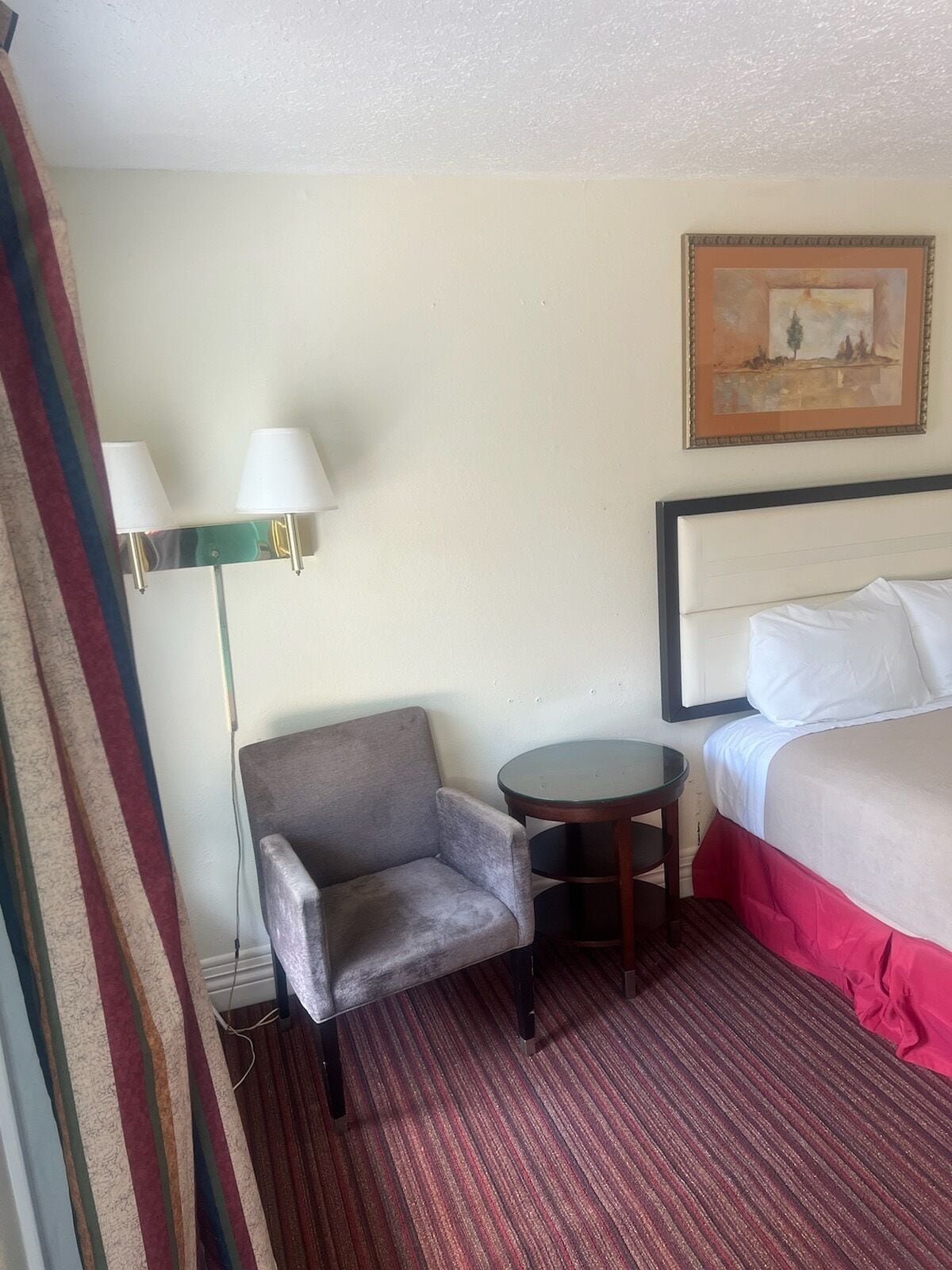Quarto de hotel com cama, uma cadeira, uma mesinha e um abajur.