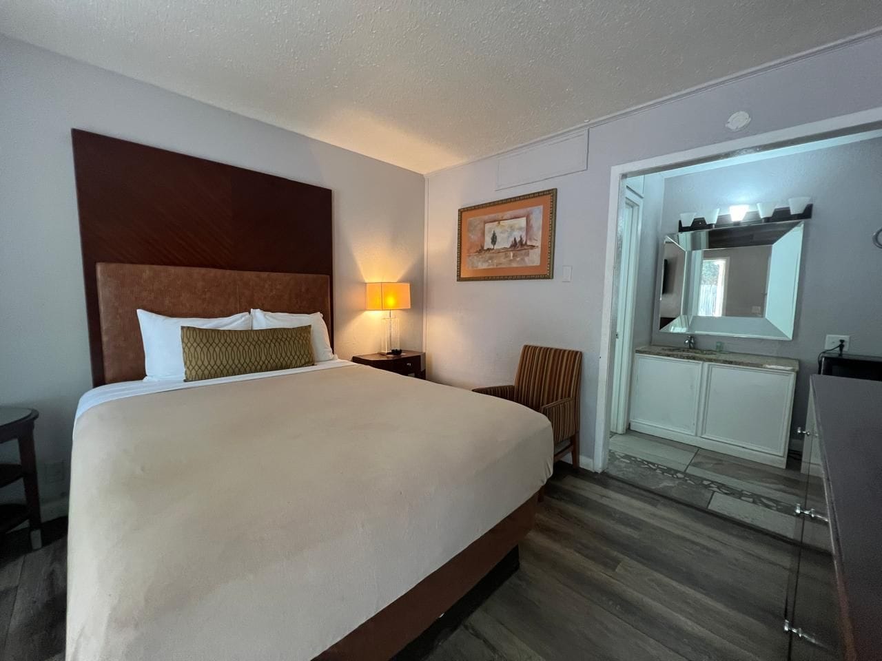 A hotel room with a large bed, a bedside lamp, a chair, a vanity with a mirror, and a small table.