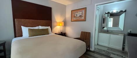 A hotel room with a large bed, a bedside lamp, a chair, a vanity with a mirror, and a small table.