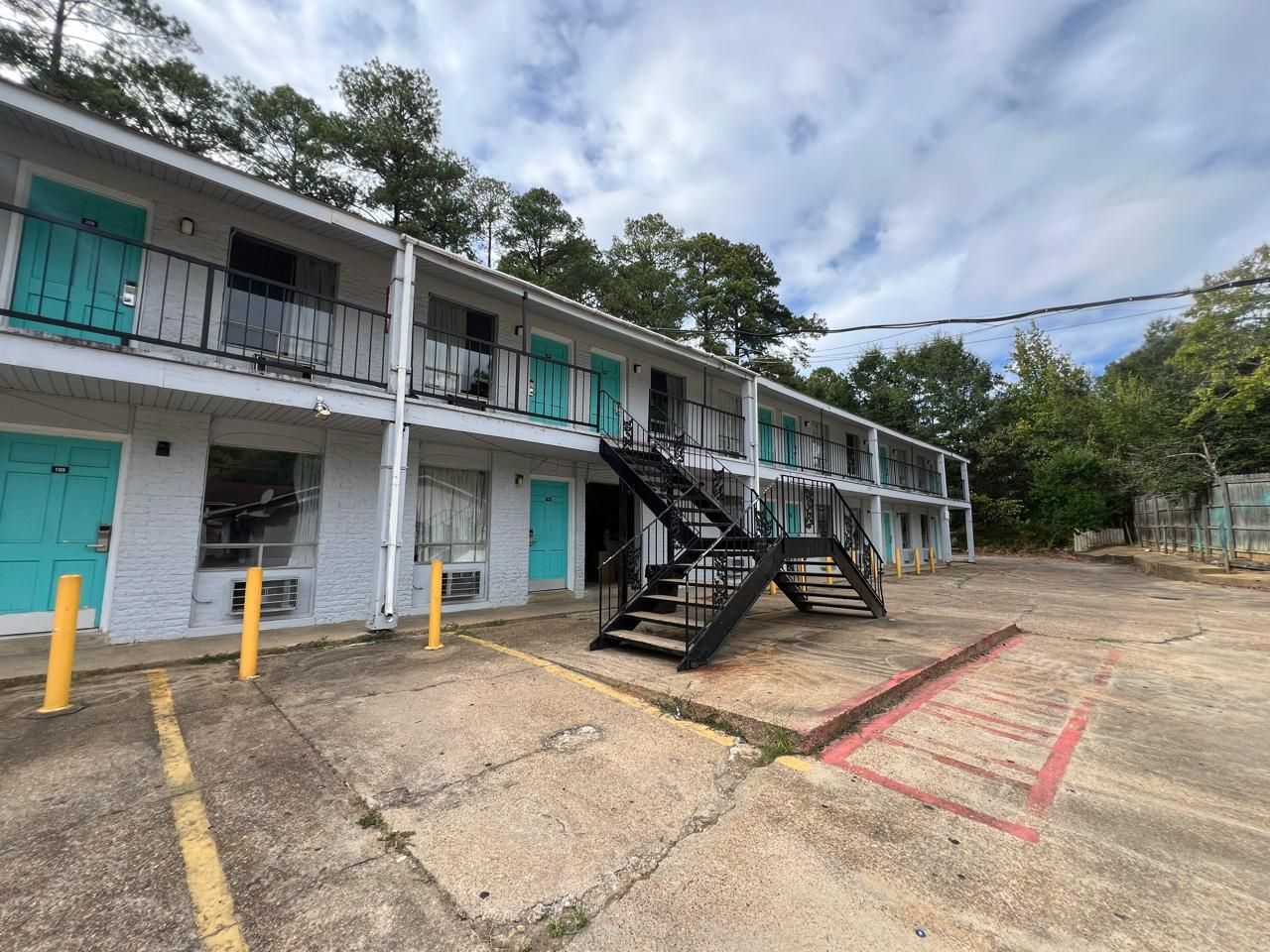 A multi-story motel with a central staircase, a parking lot with marked spaces, and a row of doors with a teal color scheme.
