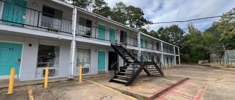 Un motel de varios pisos con una escalera central, un estacionamiento con espacios delimitados y una fila de puertas con una decoración color turquesa.