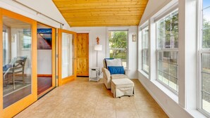 A spacious room with a wooden ceiling, large windows, a sliding glass door, a chair, a small table, and a lamp.