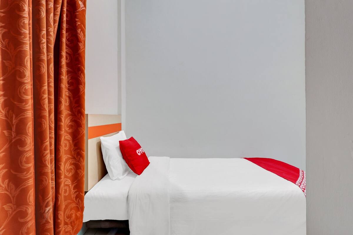 A hotel room with a neatly made bed, a red pillow, and a patterned curtain.