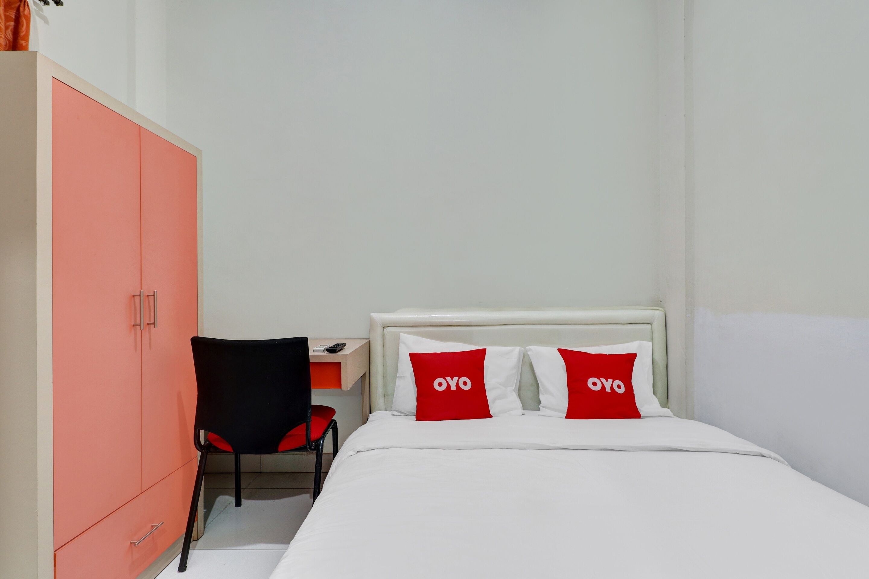 Een slaapkamer met een bed, twee rode kussens met de tekst 'OYO' erop, een wit hoofdeinde, een zwart-rode stoel en een perzikkleurige garderobe.