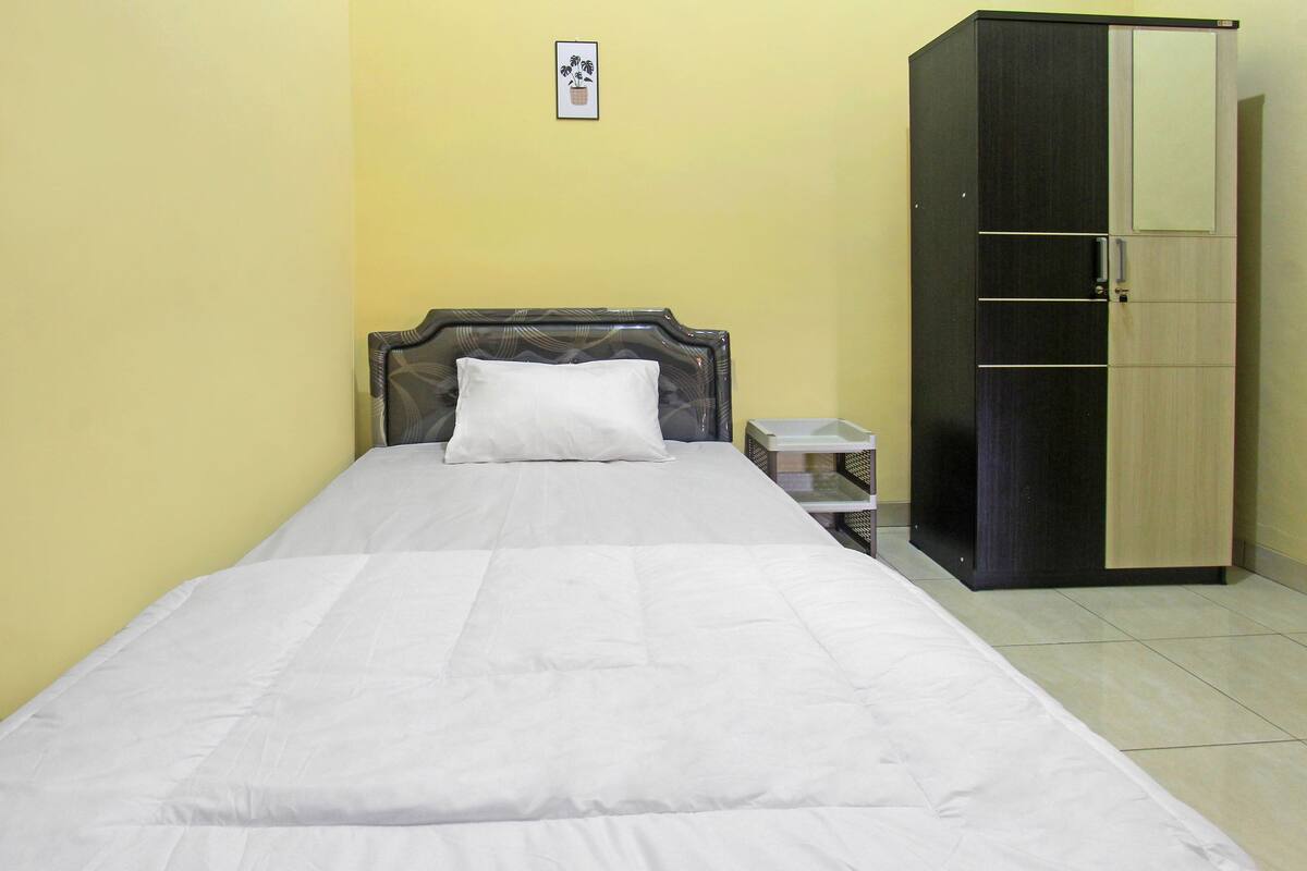 A single bed with a white mattress and a headboard, a small bedside table, and a wardrobe with a mirror.