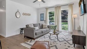 Een woonkamer met een grijze bank, een plafondventilator, een raam met gordijnen, een kleine tafel met een plant en een kamerbreed tapijt met een patroon.