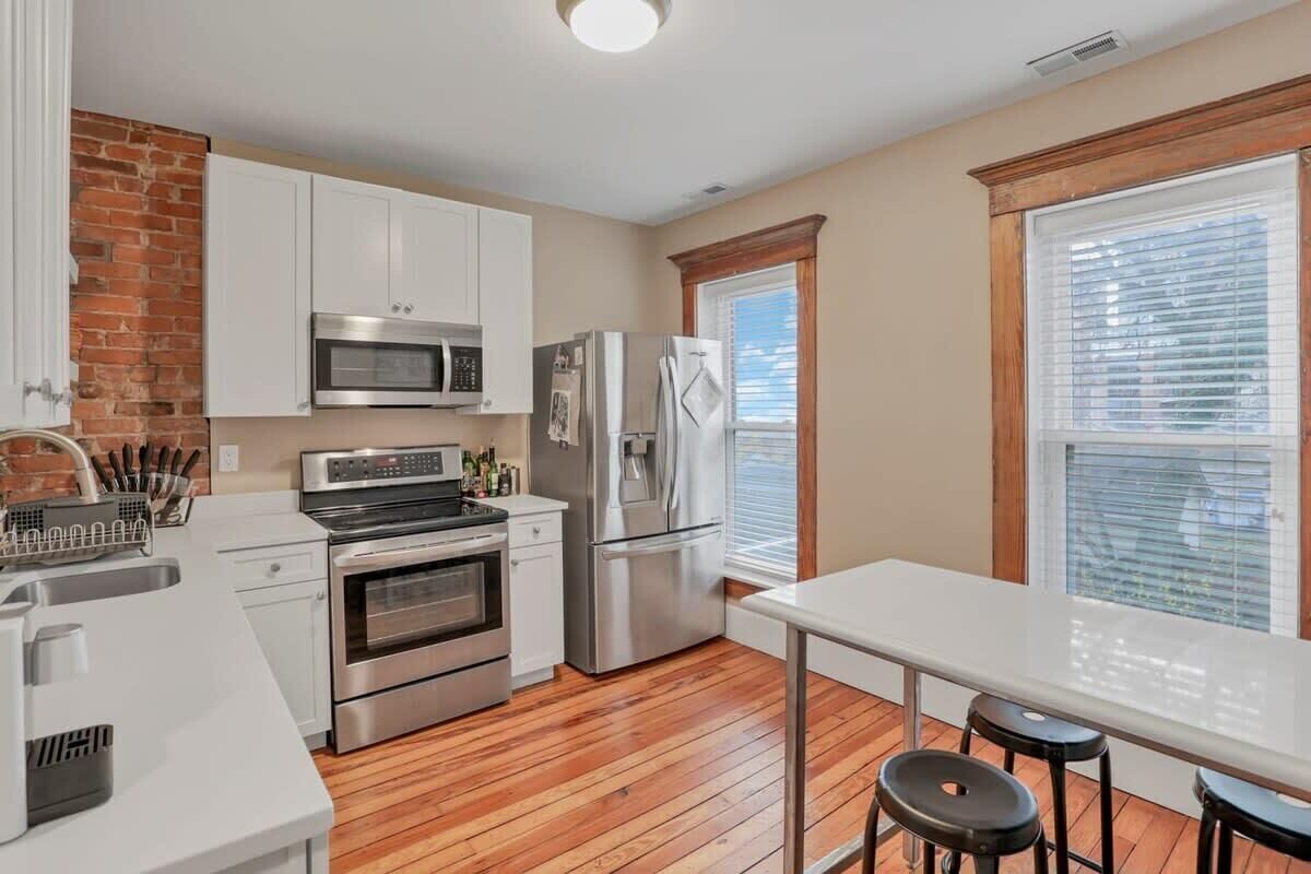 A kitchen with white cabinets, stainless steel appliances, a brick accent wall, and a window with blinds.