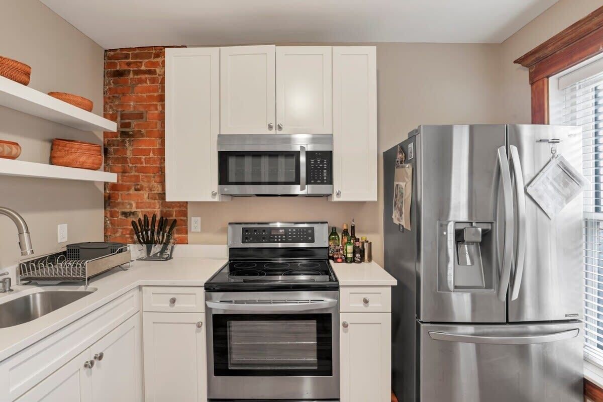 A kitchen with white cabinets, a stainless steel refrigerator, a microwave, an oven, and a brick wall.