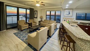 A spacious living room with a balcony, a ceiling fan, and a view of the sunset over the water.