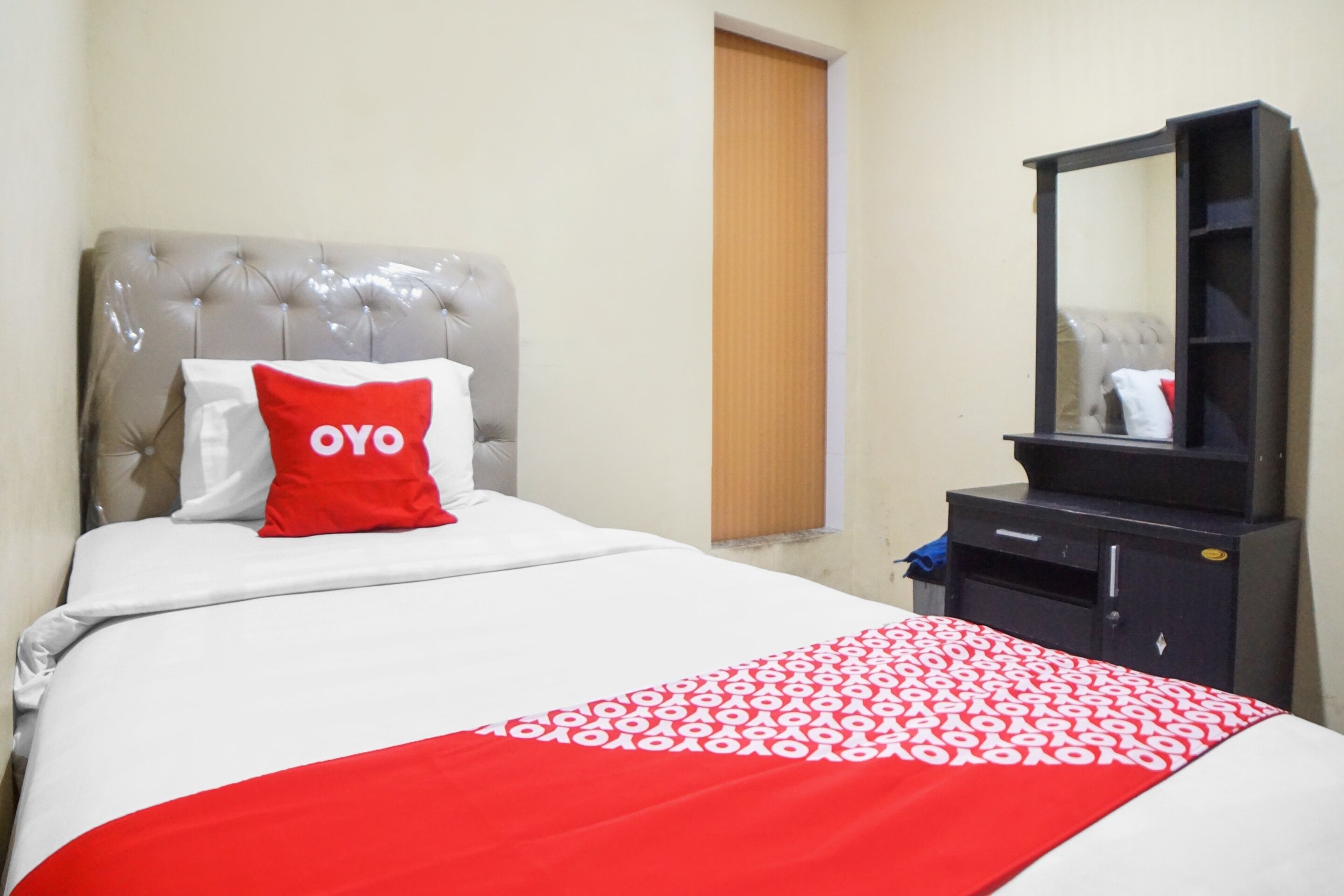 Quarto de hotel com cama, colcha estampada em vermelho e branco, cabeceira cinza com textura e cômoda preta com espelho, além de porta de madeira.