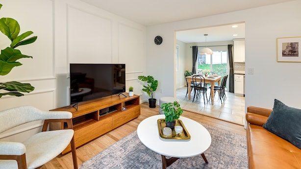 Ein modernes Wohnzimmer mit Fernseher, Holzmöbeln, einem weißen Couchtisch und einem Essbereich, der durch einen offenen Durchgang sichtbar ist.