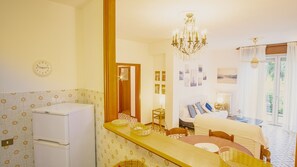 A hotel room with a bed, a dining table, a chandelier, a refrigerator, and a clock on the wall.
