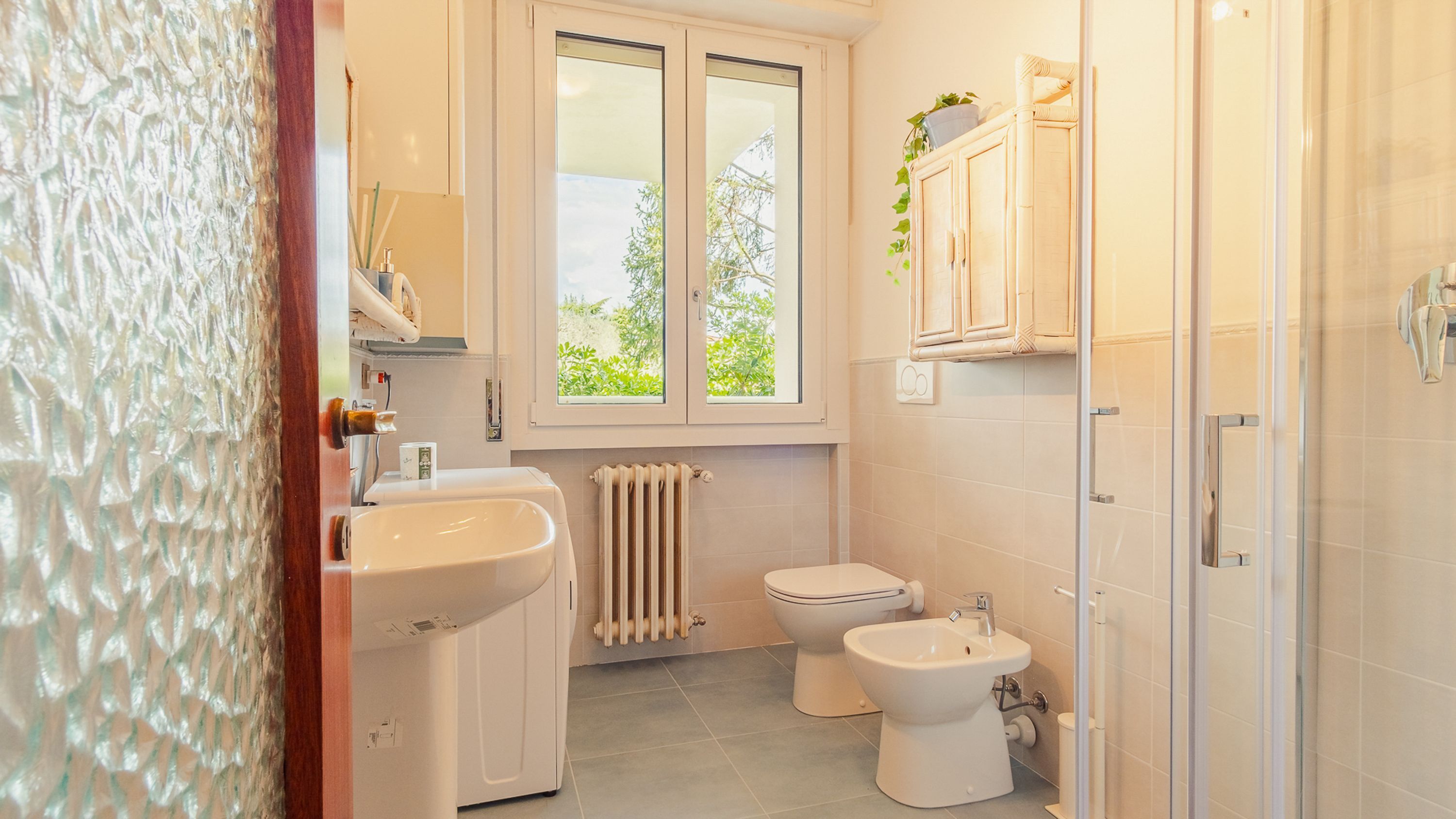 A bathroom with a toilet, bidet, sink, and a window with a view of greenery.