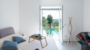 Ein modernes Schlafzimmer mit einem großen Bett, einem Nachttisch mit Büchern und einer Tasse, einem Sessel und Blick auf einen Swimmingpool und Grünfläche.