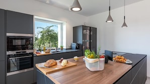 Eine moderne Küche mit dunklen Schränken, einem eingebauten Ofen, einer Kaffeemaschine und einer Holzarbeitsplatte mit frischem Obst und Brot.