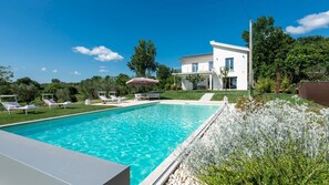 A modern house with a swimming pool, surrounded by greenery and a clear blue sky.