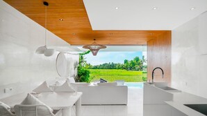 A modern kitchen with a view of a green field, white furniture, and a wooden ceiling. - Charming 1BR Villa w/ Pool | Green Flow Ubud (Ubud)