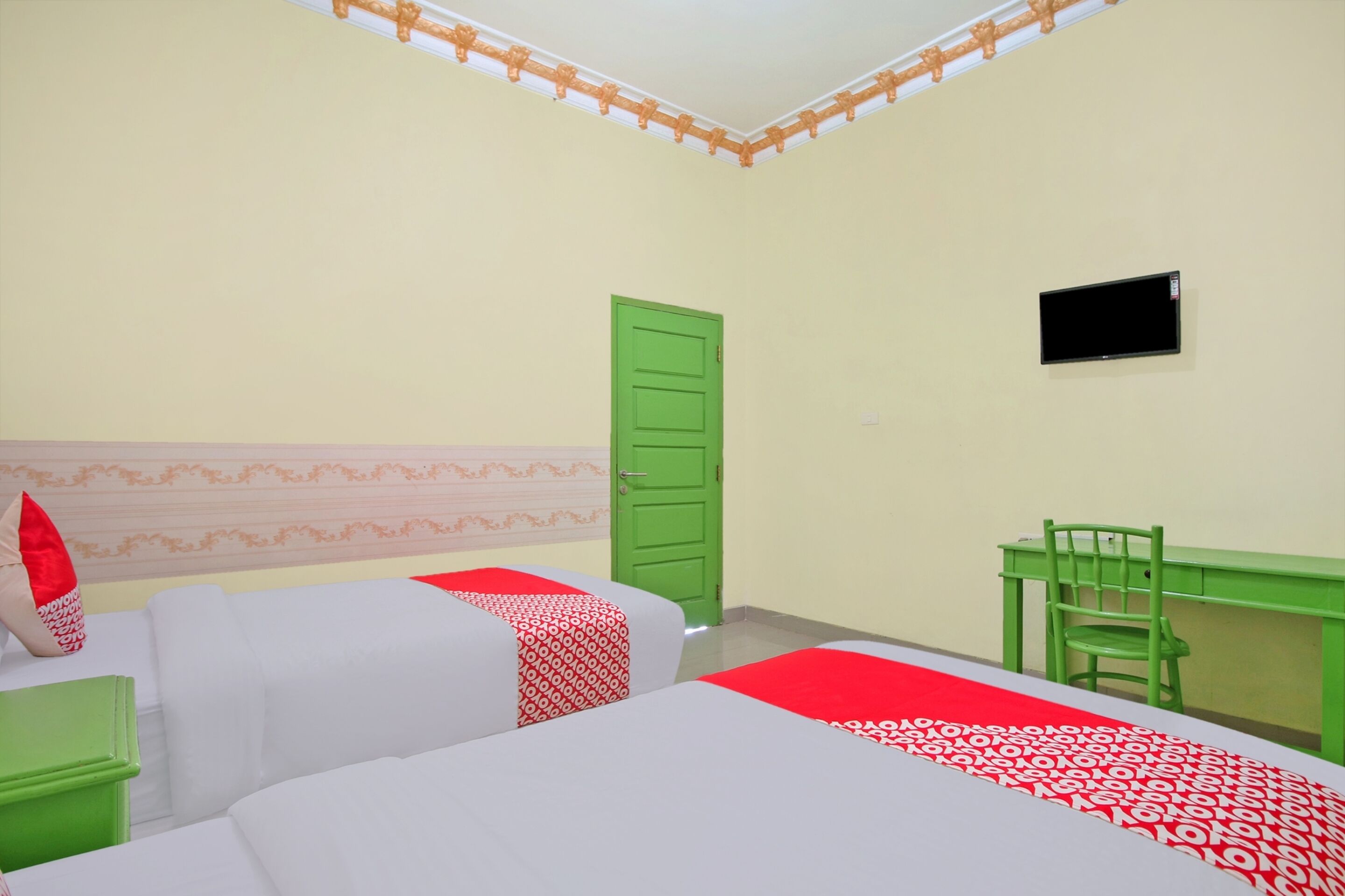Quarto com duas camas, uma escrivaninha, uma cadeira, uma televisão e uma porta verde.