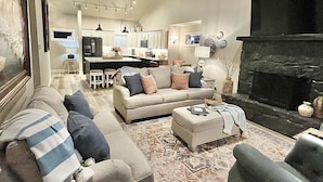 A spacious living room with a large sofa, a coffee table, and a fireplace. There is a kitchen area with a dining table and chairs in the background.