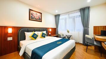 A hotel room with a bed, a desk, a chair, a small table, and a painting on the wall.