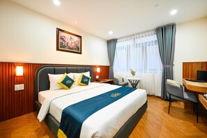 A hotel room with a bed, a desk, a chair, a small table, and a painting on the wall. - PLUS VUNG TAU HOTEL (Ho Chi Minh City)