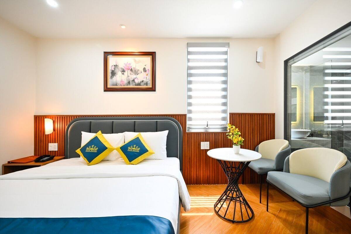 A hotel room with a bed, bedside table, lamp, artwork, window, and a small table with flowers.