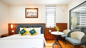 A hotel room with a bed, bedside table, lamp, artwork, window, and a small table with flowers.