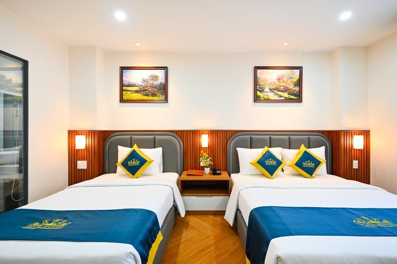 Een hotelkamer met twee bedden, elk met een houten hoofdeinde en een nachtkastje. Boven de bedden hangen twee ingelijste schilderijen.