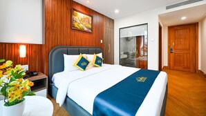 A hotel room with a bed, a nightstand, a wall-mounted painting, a bathroom door, and a wooden floor. - PLUS VUNG TAU HOTEL (Ho Chi Minh City)