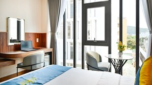Une chambre d’hôtel équipée d’un lit, d’un bureau, d’une chaise, d’un ordinateur portable et d’un balcon offrant une vue sur les bâtiments et la verdure.