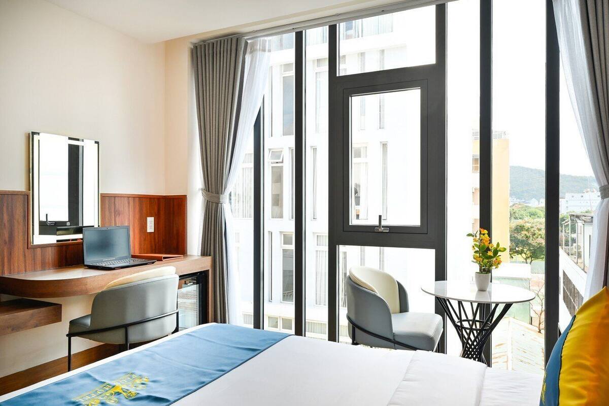 A hotel room with a bed, desk, chair, laptop, and a balcony with a view of buildings and greenery.