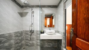 A modern bathroom with a glass-enclosed shower, a white sink, and a wooden door. - PLUS VUNG TAU HOTEL (Ho Chi Minh City)