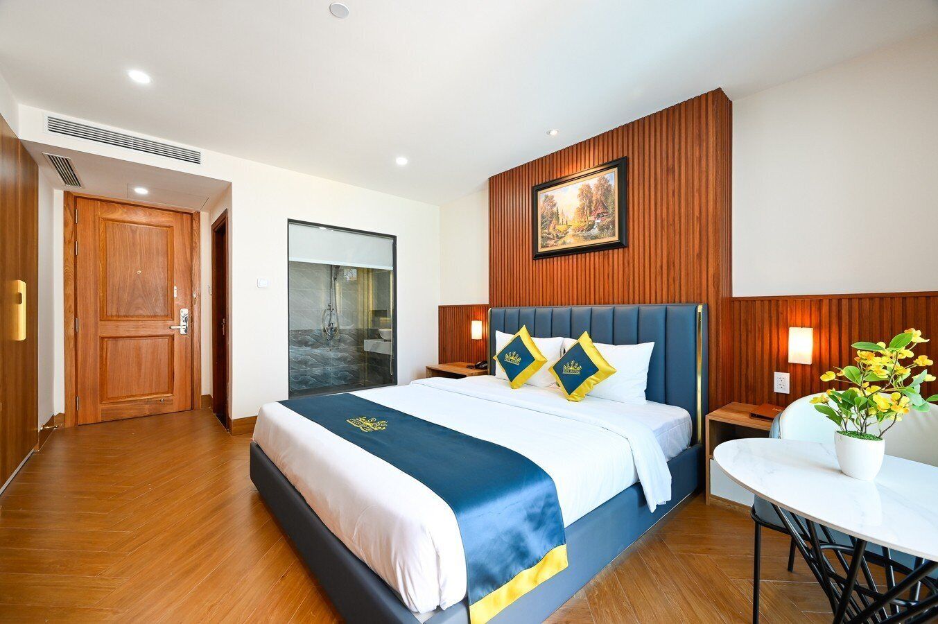 A hotel room with a large bed, wooden paneling on the walls, a painting above the bed, and a small table with a plant.