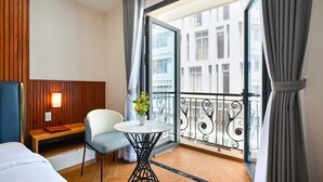 Une chambre d’hôtel comprenant un lit, un coin salon avec une table et une chaise, une plante en pot et un balcon offrant une vue sur les bâtiments.