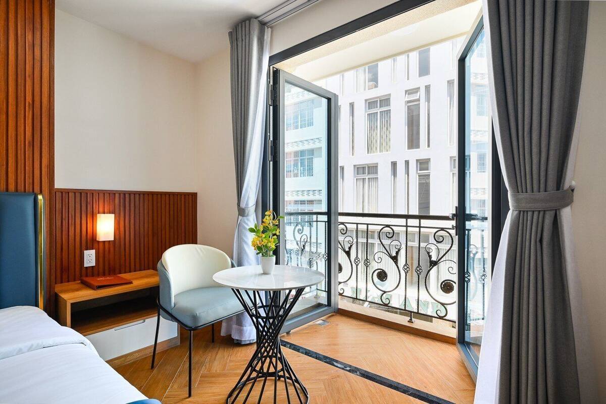 A hotel room with a bed, a seating area with a table and chair, a potted plant, and a balcony with a view of buildings.