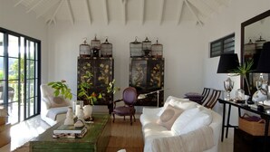 Amplia sala de estar con un sofá blanco, una mesa de centro de madera y un ventanal grande. Se observan dos aparadores de madera oscura con obras de arte, una silla y una lámpara sobre una mesita.