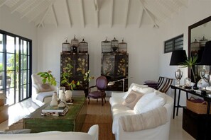 A spacious living room with a white sofa, a wooden coffee table, and a large window. There are two dark wooden cabinets with artwork, a chair, and a lamp on a side table.