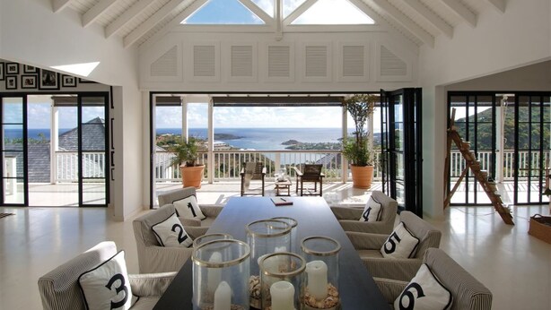 Amplia habitación con techo alto, ventanas grandes y vista al mar. Hay una mesa larga con sillas y plantas en macetas.