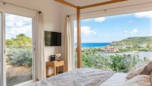 A bedroom with a large bed, a TV mounted on the wall, and a view of the sea and greenery. - Villa AU PAILLE EN QUEUE – Tranquil Escape with Ocean Views in St. Jean (Gustavia)