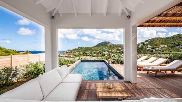 A pool area with a white sofa and lounge chairs, a wooden deck, and a view of the sea and hills. - Villa AU PAILLE EN QUEUE – Tranquil Escape with Ocean Views in St. Jean (Gustavia)