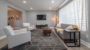 A modern living room with a grey sofa, a grey armchair, a wooden coffee table, and a TV mounted on the wall. - Koselig Retreat (Randallstown)