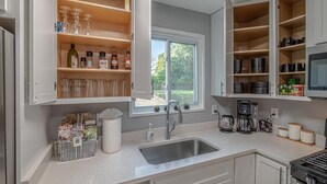 A kitchen with open cabinets, a window, and a stainless steel sink. - Koselig Retreat (Randallstown)