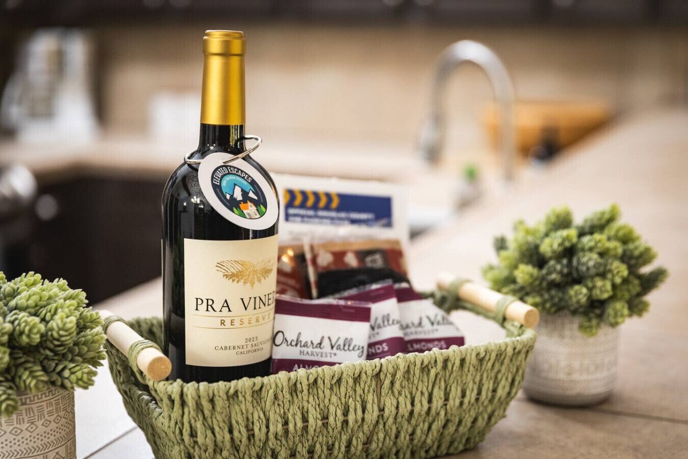 Een fles Pra Viner Reserve 2023 Cabernet Sauvignon in een gevlochten mand met twee kleine potplanten en een doos Orchard Valley Harvest wijn.