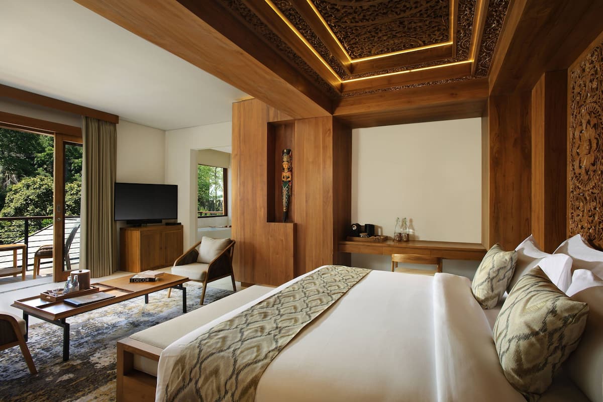 A hotel room with a large bed, a desk, a TV, and a balcony with a view of trees.