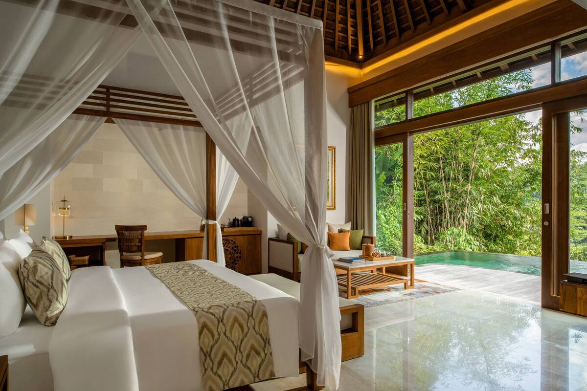A spacious bedroom with a large bed, a wooden desk, and a view of lush greenery through large windows.