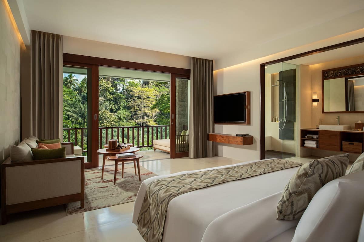 A modern hotel room with a large bed, a TV, a bathroom with a mirror, and a balcony with a view of the greenery.