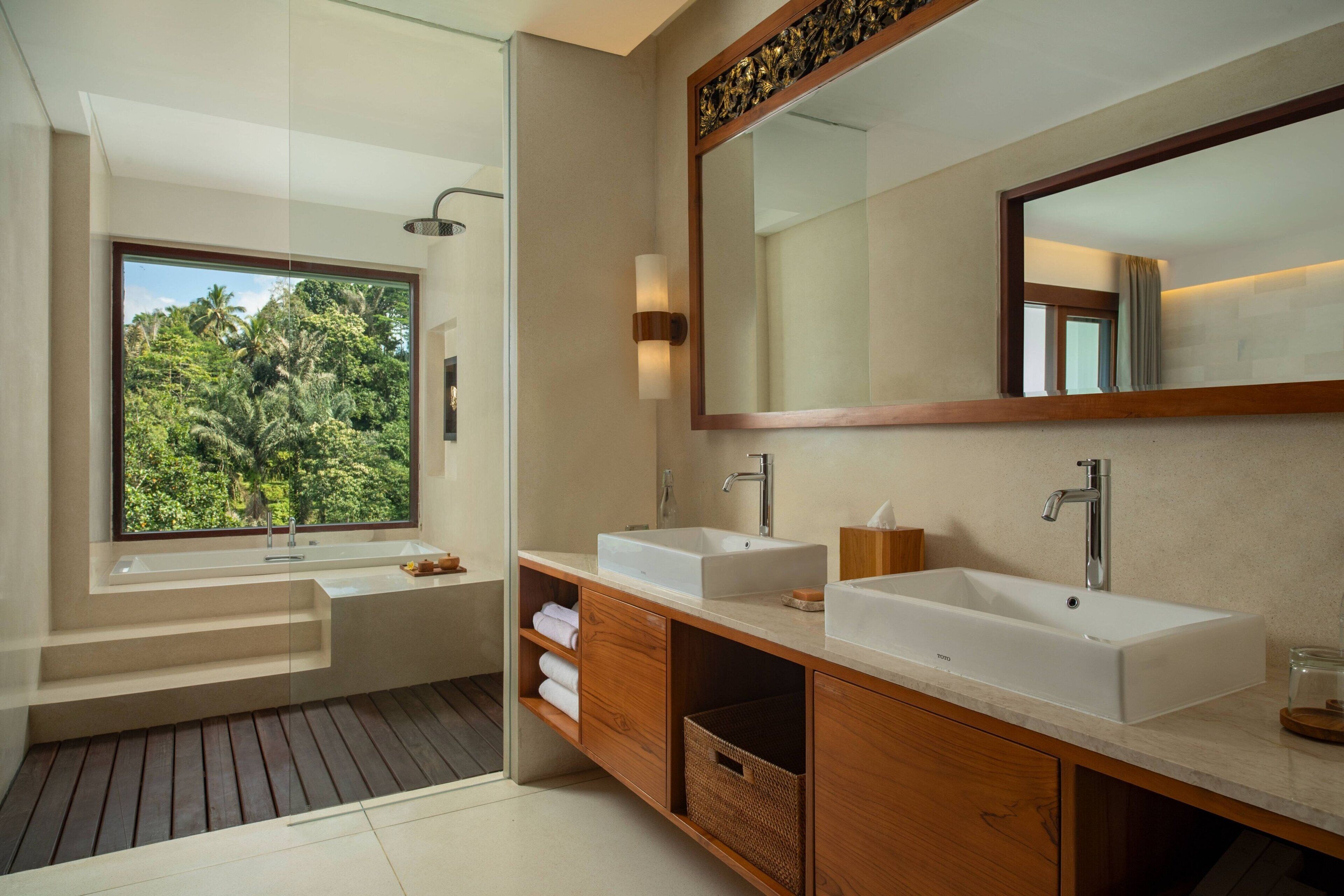 A modern bathroom with two sinks, a large mirror, and a window offering a view of lush greenery.