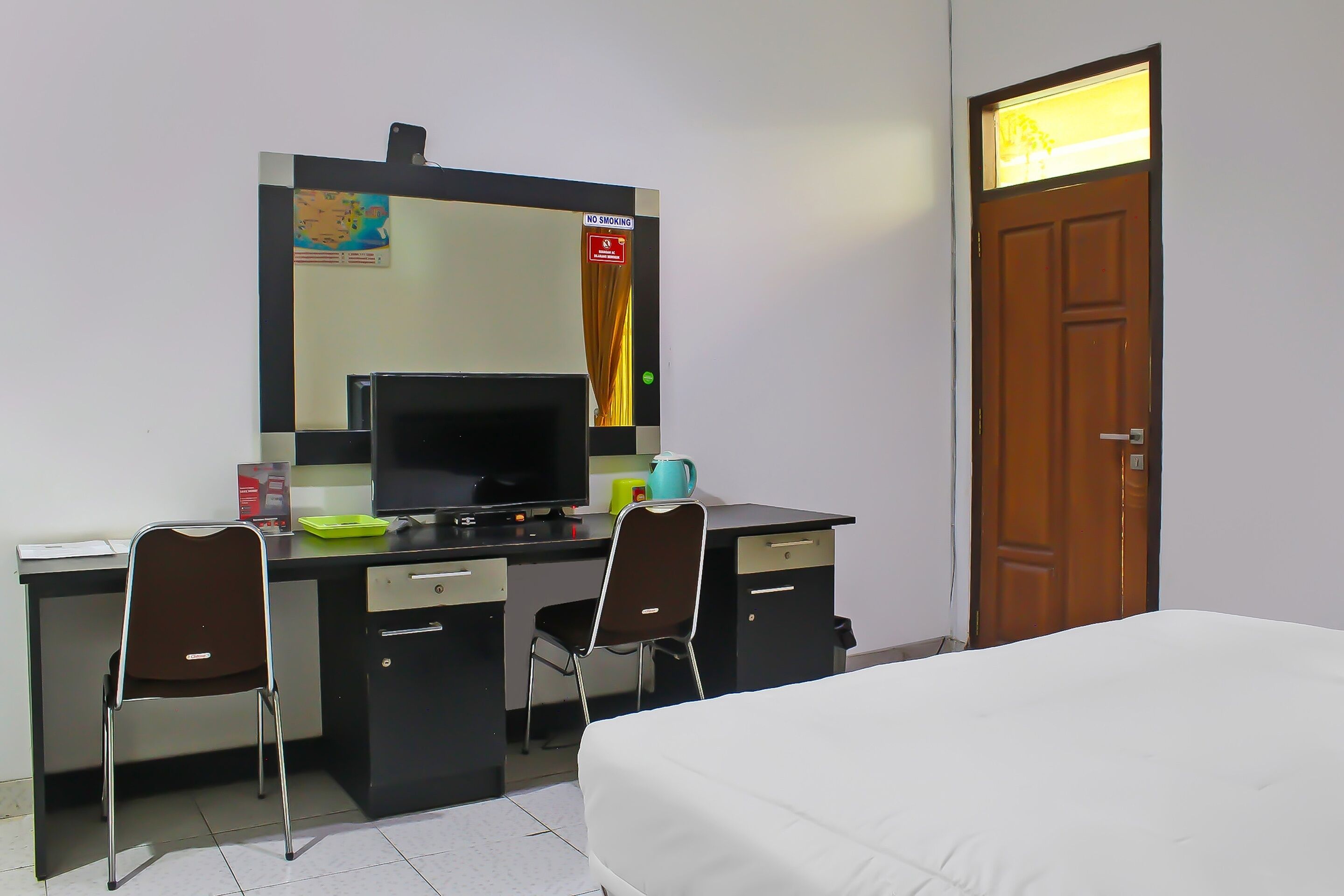 A hotel room with a bed, a desk with a computer, two chairs, a wooden door, and a mirror.